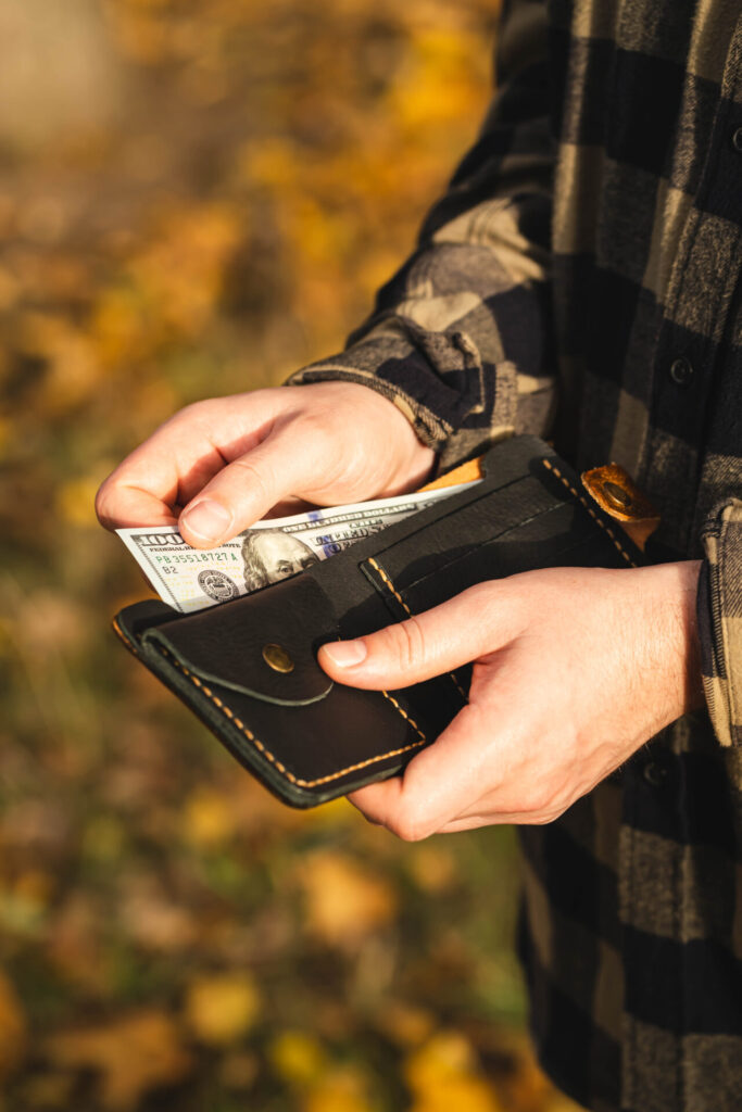 Person in a checkered shirt holds an open black wallet with visible hundred-dollar bills. The background has a warm, autumnal tone with blurred foliage.