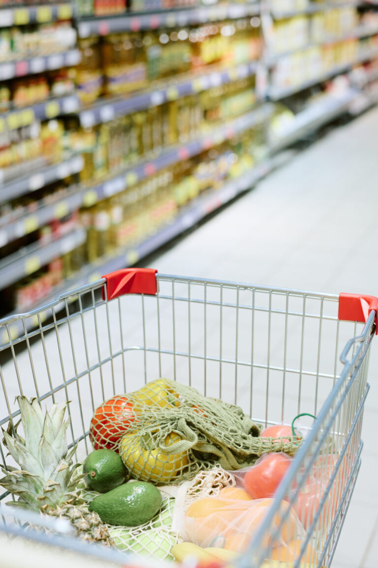 Fresh produce in a shopping cart, featuring pineapple, avocado, and oranges, displayed in a supermarket aisle.