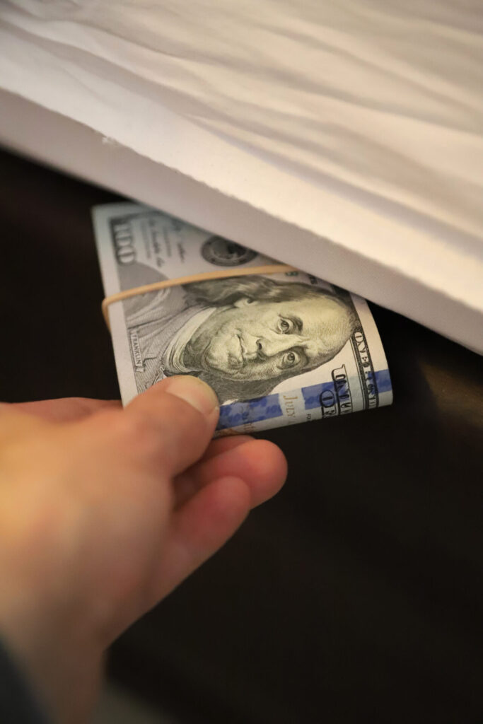 Person hiding a wad of dollar bills under a mattress, indicating a secretive approach to saving money at home.