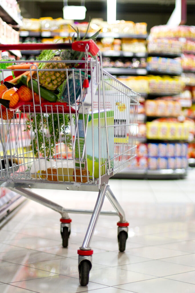Shopping cart in a supermarket filled with fresh groceries, including fruits, vegetables, and packaged items.